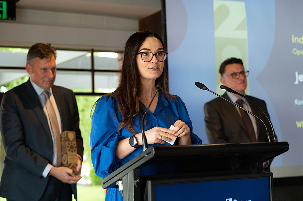 Jessica Bobbinmarsilio standing at lectern giving a speech with two people standing behind them, one holding a trophy.
