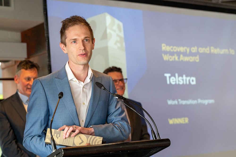 Tim Saunders standing at lectern giving a speech with trophy in hand standing in front of a screen with writing ‘Recovery at and Return to Work Award. Telstra. Work Transition Program. Winner’.